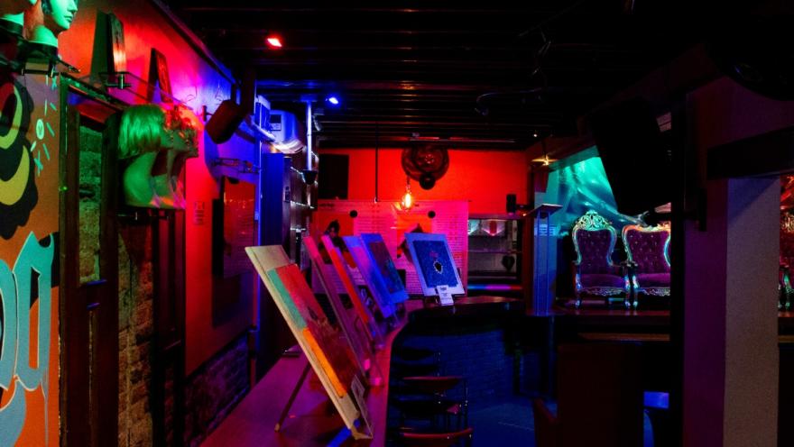  Museo LGBTI sala oscura con paredes con adornos de colores y fotografías. Se ven 3 cabezas de maniquies y dos sillas clásicas.