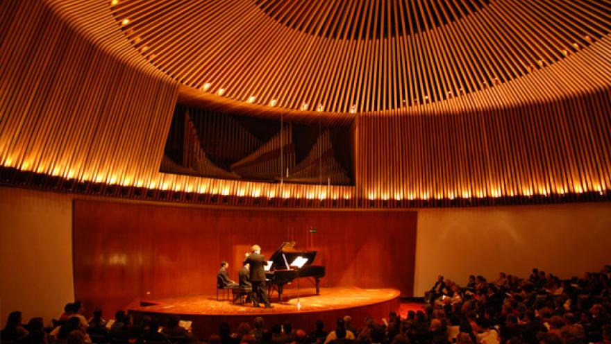 Biblioteca Luis Ángel Arango, al interior Sala de conciertos con músicos en tarima tocando un piano de cola café. El salón con paredes y techos de madera de un color rojizo está repleto. 