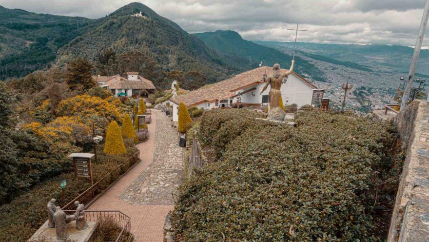 Monserrate Hill, inner pathway lined with medium-sized pines and trees, featuring bronze statues, two buildings, and in the background, Cerro de Guadalupe and part of the city’s southwest.