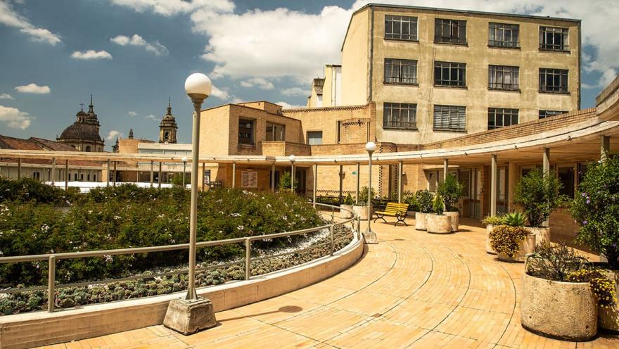 La Candelaria - Centro Gabriel Garcia Marquez, un corredor circular en ladrillo en pisos y paredes con materas, sillas y faroles de luz alrededor. Se destaca atrás un edificio antiguo y al fondo las cúpulas de la Catedral Primada.