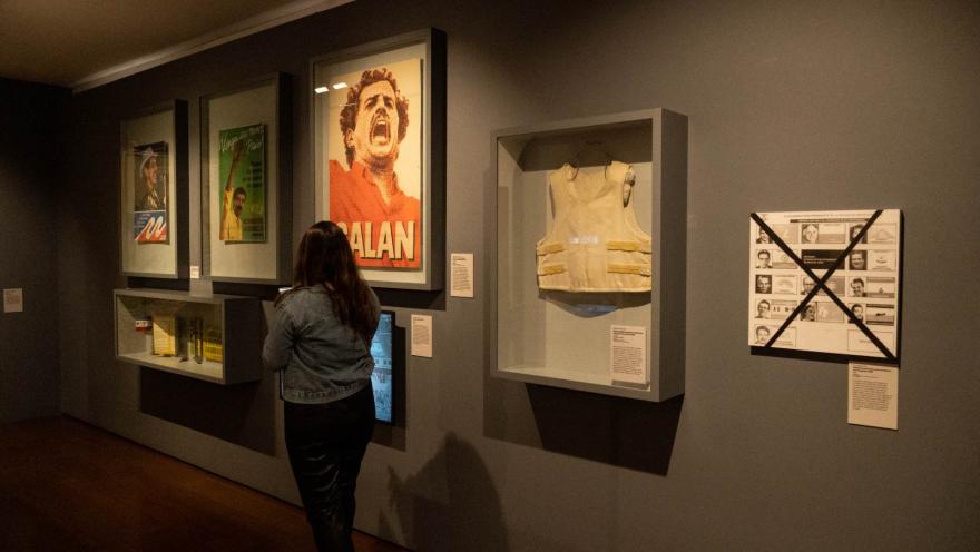 Museo Nacional de Colombia al interior salón con exhibición de imágenes de lideres políticos desaparecidos víctimas de la violencia como Luis Carlos Galán, Carlos Pizarro y Bernardo Jaramillo. Hay una vitrina con un chaleco antibalas y una mujer de espaldas observando las obras.
