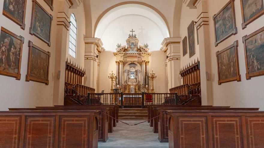 Catedral Primada de Colombia, capilla interior con dos paredes repletas de cuadros, dos filas de sillas en madera y al fondo un altar dividido por una reja metálica. El salón está vacío.