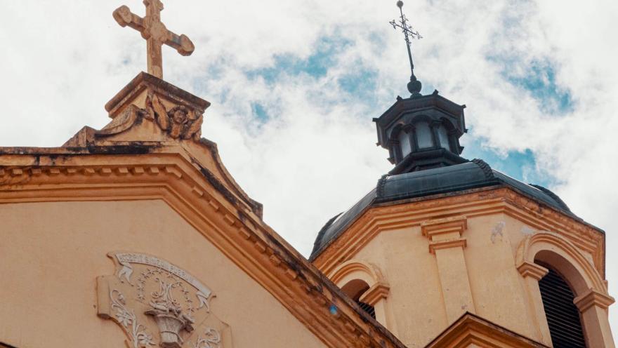  Iglesia de la Candelaria Iglesia de la Candelaria colonial con la parte alta de su fachada en primer plano, se resalta la punta del frente coronada por una cruz y una torre con su cúpula negra y una cruz metálica encima. 