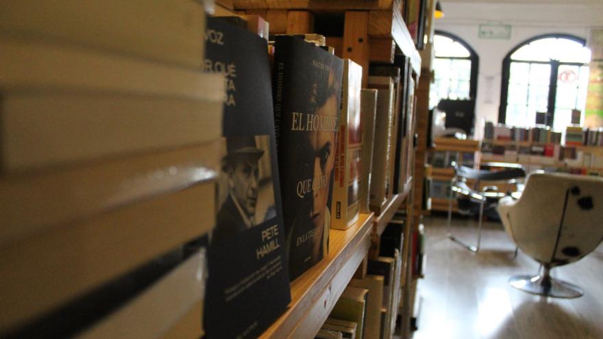 Garabato Libros primer plano en lateral a la izquierda de libros puestos sobre estantería de madera. Se alcanza a leer un título sobre Sinatra. Al fondo se observan un par de sillas metálicas, una estantería difusa y unos ventanales con luz hacia la calle.