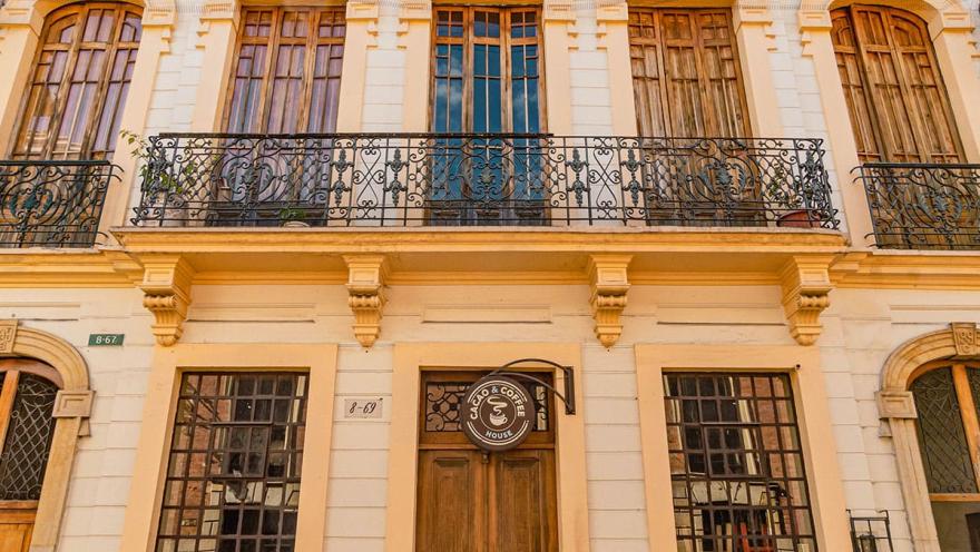 La Candelaria fachada edificio republicano con cinco ventanas en madera en el segundo piso, balcón amplio con rejas metálicas, y en el primer piso una puerta grande y dos ventanales. Un letrero redondo y pequeño dice Cacao & Coffee House.