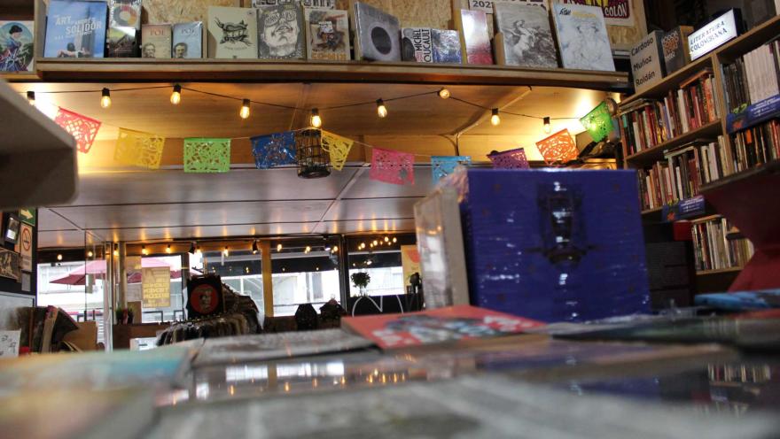 Librería La Valija de Fuego con libros en una mesa y al fondo un salón iluminado con una manguera de varios bombillos con luz amarilla y libros puestos en un anaquel en una pared cerca al techo. Al fondo se ve un ventanal hacia la calle.