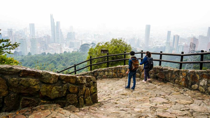 Sendero Cerro Monserrate en el camino de piedra con un hombre con bastón y una mujer caminando. Detrás de la barrera de madera se ve la panorámica de la ciudad entre nubes espesas.