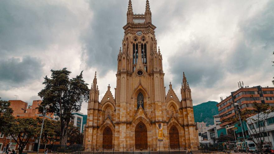 Iglesia Lourdes plano general de frente a la fachada gótica. Las 3 puertas están cerradas y la proteje una reja metálica negra. Al lado derecho hay varios árboles, uno de ellos muy alto. Al frente en la plazoleta camina una mujer, hay varias palomas en el suelo y un grupo de personas está sentado bajo un arbol. El cielo está gris.