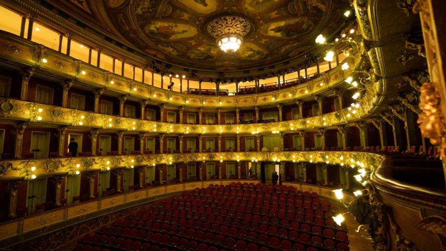 Teatro Colón interior, wide general shot from a balcony above the stage, with a view of the last row of seats and the access door where a person is standing. The rest of the venue is empty, with the lights on.