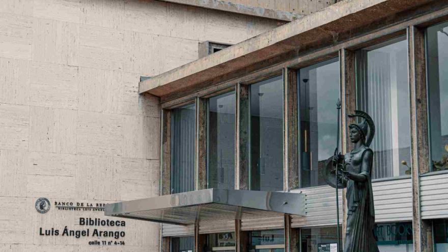 Biblioteca Luis Ángel Arango en primer plano de la entrada principal donde se la puerta en vidrio, una estata a la izquierda y a la derecha en la pared el aviso del nombre del lugar.