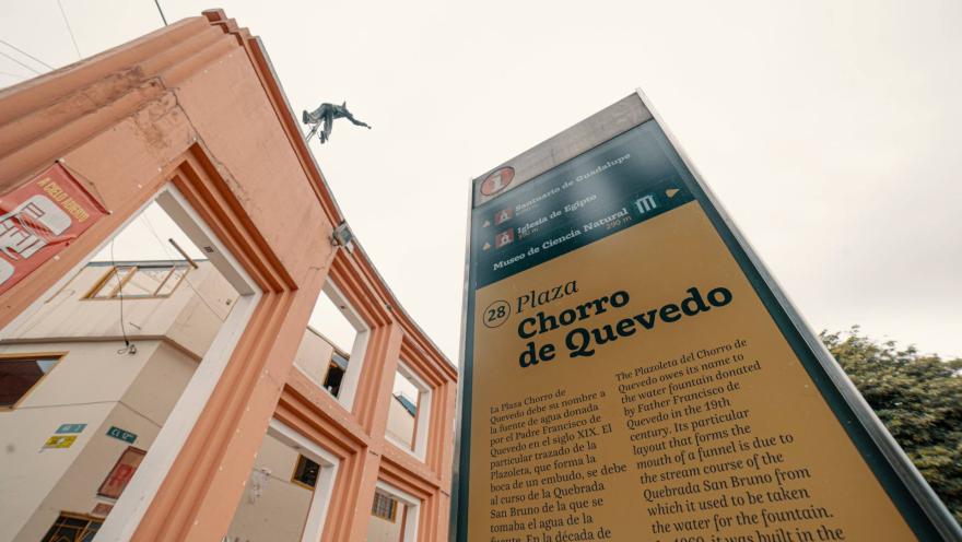 Plazoleta del Chorro de Quevedo en plano contrapicado de señal turística, pared y escultura de payaso en monociclo. El cielo es blanco.