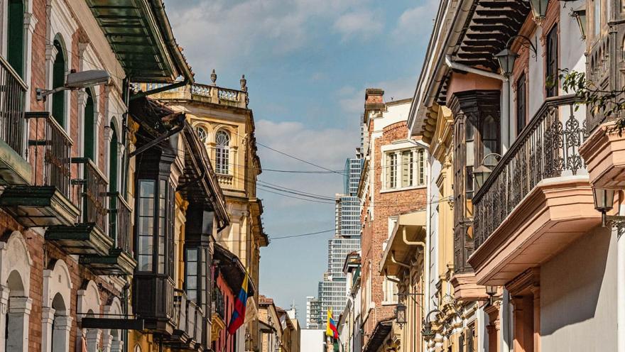 La Candelaria vista calle colonial con casas con balcones.. Hay un pare de banderas de Colombia y al fondo se ve un edificio moderno, el Bacatá.
