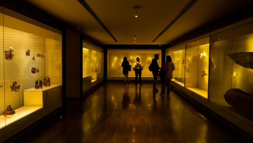 Museo del Oro en salón de exhibición con 6 vitrinas que exhiben piezas, que son observadas por 4 personas.