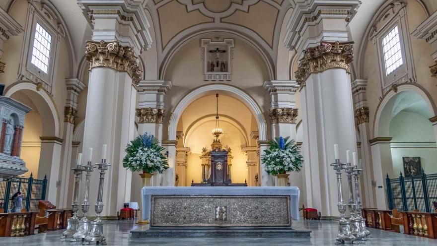 Catedral Primada de Colombia, en plano general del atrio en forma de arco con sus 3 columnas a cada lado, 3 candelabros grandes con sus velones a cada lado, 2 arreglos florales a cada lado, presidido arriba por un cristo en la cruz y al fondo un retablo del Santísimo iluminado con una lámpara de luz amarilla.