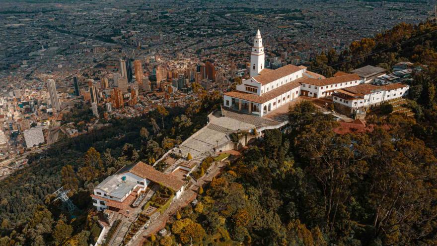 Sendero Cerro Monserrate en plano desde arriba de la iglesia en la cima montaña con árboles frondosos alrededor y la ciudad con edificios altos de fondo.