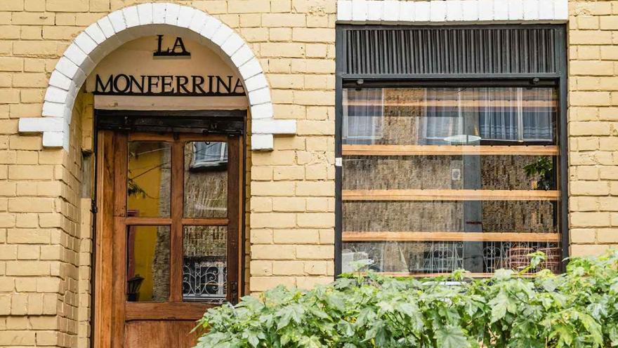Esta fotografía de cerca muestra la fachada de "LA MONFERRINA", un restaurante en el barrio de La Macarena. La arquitectura tradicional del lugar se destaca por su fachada de ladrillo amarillo y una puerta de madera de estilo rústico enmarcada por un arco blanco. El toque de naturaleza lo da un seto de plantas verdes en la parte inferior, que le da vida y color al lugar.
