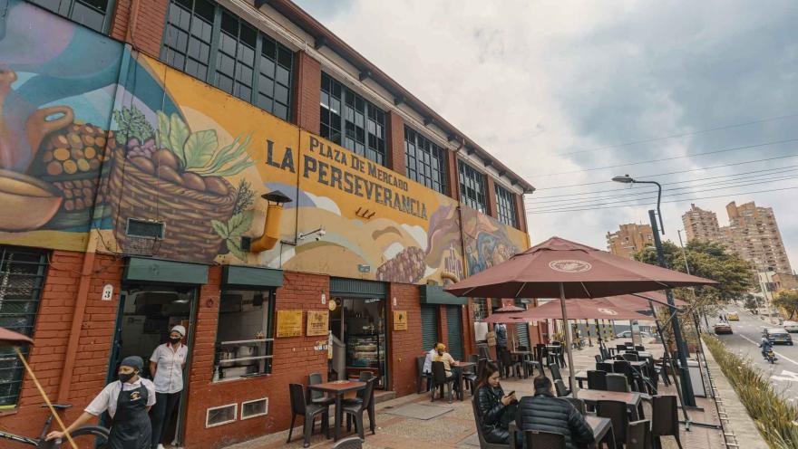 Esta es una vista exterior de la "Plaza de Mercado La Perseverancia", un edificio de ladrillo rojo con grandes ventanas en el piso superior. La fachada está adornada con un vibrante mural que representa escenas de mercado y paisajes. Delante del edificio, hay una zona de mesas y sillas con sombrillas de color marrón, donde varias personas están sentadas. En el lado izquierdo, dos personas con mascarillas, una de ellas con un delantal, están en la entrada. A la derecha, se ve una calle con tráfico y edificio