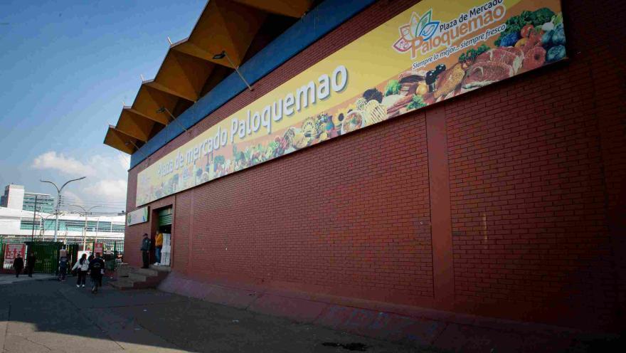  La imagen muestra la fachada exterior de la "Plaza de Mercado Paloquemao" bajo un cielo despejado. El edificio tiene una estructura de ladrillo rojo y una marquesina distintiva con un patrón en zigzag en la parte superior. Un gran letrero amarillo horizontal exhibe el nombre del mercado y un colorido mural con ilustraciones de frutas, verduras, carnes y pescados, junto con el eslogan "Siempre lo mejor, siempre fresco". Varias personas se ven entrando y saliendo de la entrada principal a la izquierda.