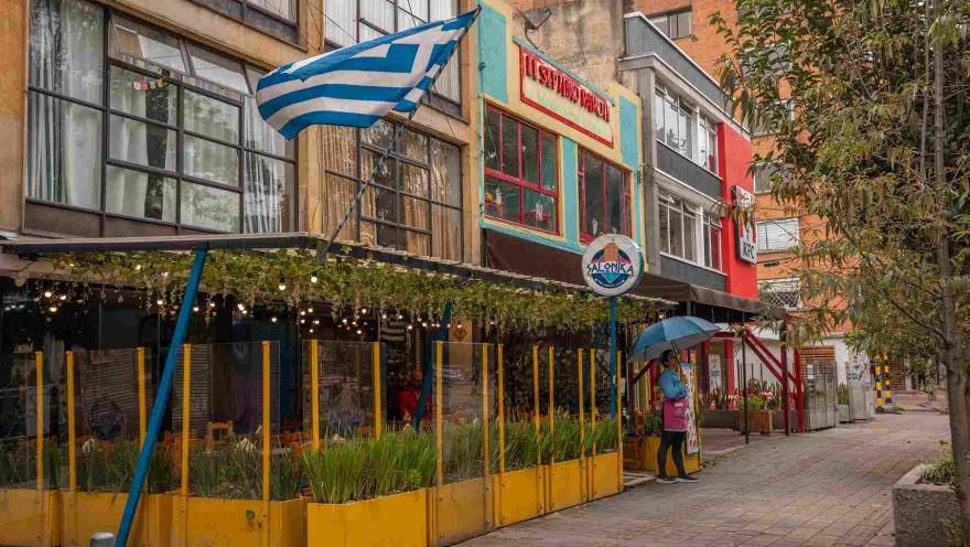 Esta imagen muestra una calle vibrante y llena de color en el Park Way. En primer plano, la fachada de un restaurante de comida griega llamado 'Salonika´ se destaca por sus colores alegres. Un toldo de metal y vidrio con luces colgantes cubre la terraza, creando un ambiente acogedor. La bandera griega ondeando sobre el restaurante y los detalles en amarillo y azul evocan la cultura mediterránea.