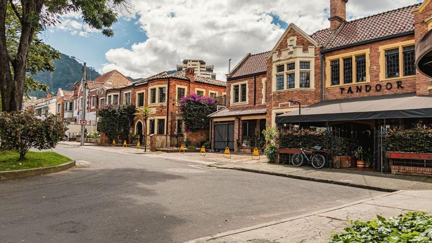 Esta imagen captura la esencia de la Quinta Camacho, un barrio de Bogotá caracterizado por su arquitectura de estilo inglés. Se aprecian varias casas de ladrillo rojo, con techos a dos aguas y ventanas enmarcadas, que se alinean a lo largo de una calle tranquila y arbolada. El ambiente es sereno y residencial, con un cielo parcialmente nublado que realza los colores de las fachadas y la vegetación.