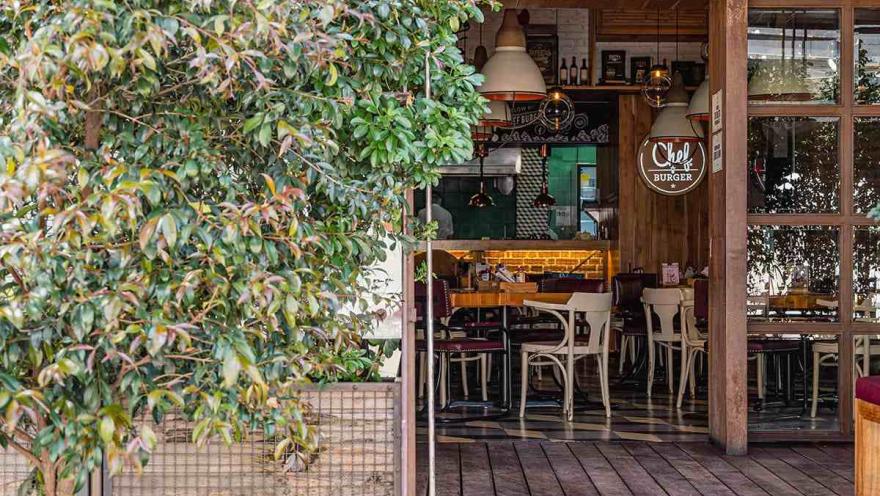 La fotografía muestra la entrada de un restaurante llamado "Chef Burger" en el Parque de la 93. La arquitectura del lugar es moderna y acogedora, con un exterior de madera y grandes ventanales que permiten ver el interior. Las lámparas de estilo industrial cuelgan sobre las mesas, creando un ambiente cálido e íntimo. El diseño del espacio invita a los transeúntes a entrar y disfrutar de una comida en un ambiente relajado y contemporáneo.