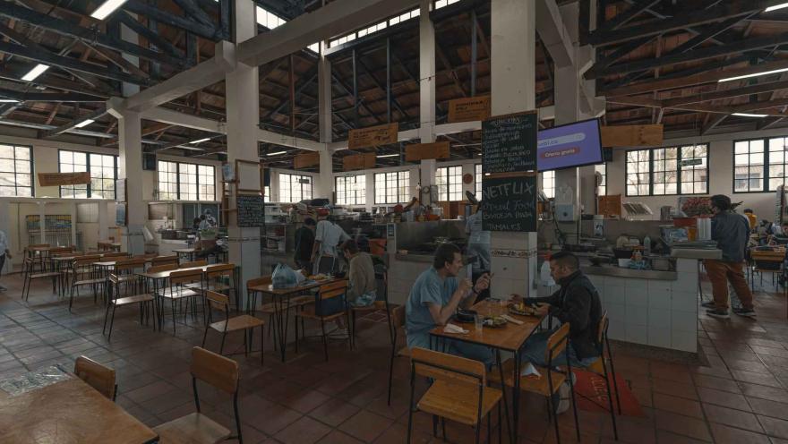 La imagen muestra el interior espacioso de una plaza de mercado o un patio de comidas. Varias personas están sentadas en mesas de madera y sillas de metal, disfrutando de sus comidas. Al fondo, se pueden ver diferentes puestos de comida con cocineros y personal atendiendo. El techo alto, con vigas de madera y metal, y las grandes ventanas, sugieren una estructura antigua y bien iluminada. Un televisor montado en la pared y letreros escritos a mano añaden detalles al ambiente.