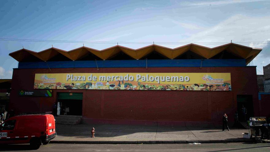 Esta imagen presenta una vista frontal más amplia de la fachada de la "Plaza de Mercado Paloquemao". El edificio de ladrillo rojo está coronado por una marquesina de techo con un diseño ondulado y escalonado en tonos amarillos y azules. Un gran letrero amarillo con el nombre del mercado y un mural ilustrado de productos frescos se extiende a lo largo de la pared. En primer plano, se ve una acera con un hidrante rojo y algunas personas caminando. Un vehículo rojo está estacionado a la izquierda.