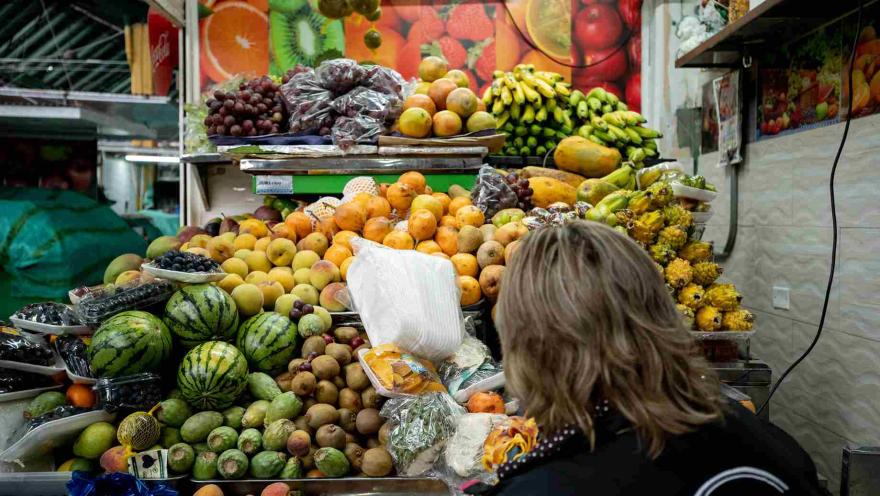 Esta imagen se centra en una vibrante exhibición de frutas frescas en un puesto de mercado. Una persona con cabello largo y rubio, vestida de negro, se encuentra de espaldas, mirando la gran variedad de frutas. Melones, uvas, plátanos, mangos, papayas y otras frutas exóticas están apiladas en abundancia. Un mural de frutas se ve en el fondo.