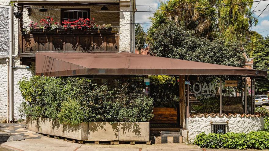 Esta foto de cerca muestra la fachada de Rose. La estética del lugar es moderna y chic, dominada por un color rosa pastel y detalles en blanco. A través de los grandes ventanales, se ven las decoraciones interiores con toques tropicales, como hojas de palmera blancas que contrastan con el interior minimalista. El diseño del espacio es un claro ejemplo de la renovación que ha experimentado el barrio.