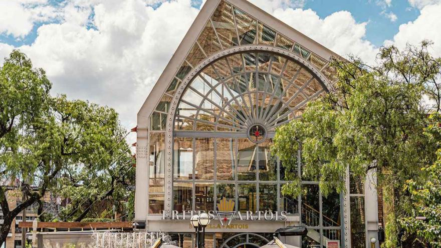 Esta fotografía muestra la imponente arquitectura de un edificio en la Zona G, que alberga un restaurante llamado 'Frites Artois'. La estructura se caracteriza por su gran fachada de vidrio en forma de arco, con detalles de metal que le dan un aire industrial y moderno. El edificio está rodeado de árboles frondosos y, en primer plano, se ven las sombrillas de las terrazas de los restaurantes, que invitan a los comensales a disfrutar del aire libre.