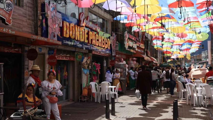 Esta fotografía se centra en la calle principal, donde las sombrillas de colores crean un toldo flotante, añadiendo un toque festivo al ambiente. El letrero prominente de "DONDE PACHECO" sugiere la presencia de un reconocido negocio de lechona o comida tradicional colombiana. La calle está llena de gente, algunos sentados en mesas de plástico blanco disfrutando de la comida, y otros paseando por la acera. La escena evoca un día soleado y animado, donde la gastronomía y la cultura se mezclan
