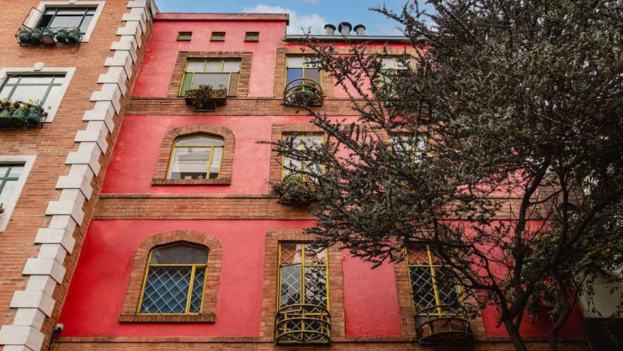 Esta foto de una fachada en La Macarena resalta la combinación de estilos y el uso del color. El edificio, con su llamativa fachada de color rojo con detalles en amarillo, se ve en contraste con un edificio de ladrillo en el lado izquierdo. Las ventanas con rejas, los balcones circulares y los adornos en la parte superior le dan un toque único y llamativo. La textura de la pintura y los detalles de la arquitectura se aprecian con claridad, lo que resalta el carácter artístico de la zona.
