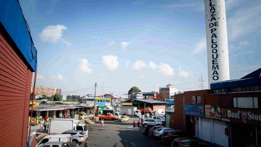 La fotografía ofrece una vista elevada de un área interior concurrida de la "Plaza de Mercado Paloquemao". Una calle asfaltada atraviesa el centro, con varios coches estacionados y personas caminando. A la izquierda, se observan puestos de mercado al aire libre con toldos. A la derecha, un edificio de ladrillo y una alta columna blanca con el texto "PLAZA DE PALOQUEMAO" escrito verticalmente. El cielo azul con algunas nubes dispersas se extiende por encima de un paisaje urbano.