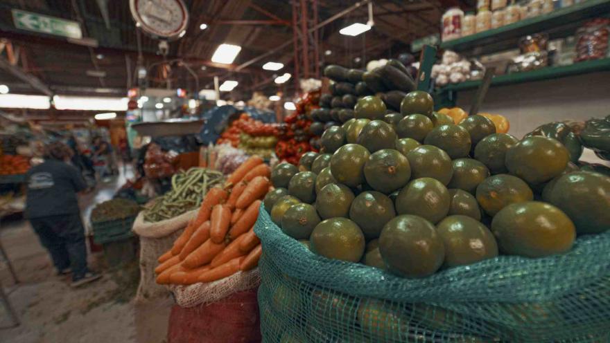 En esta imagen, un primer plano enfocado en grandes sacos de frutas cítricas verdes domina el lado derecho. Detrás y a la izquierda, se ven sacos de zanahorias, calabacines y otros vegetales apilados. Al fondo, se distingue el ambiente bullicioso de un mercado, con una persona caminando y estantes llenos de productos. Una báscula circular se ve desenfocada en la parte superior izquierda.