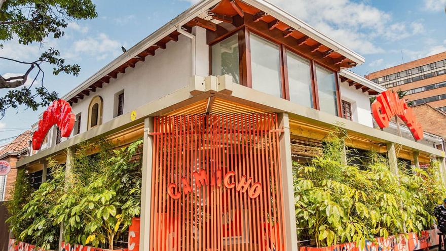  La imagen presenta la fachada de Camacho, un restaurante con un diseño llamativo y contemporáneo. El edificio, de color blanco y con toques de madera, se destaca por su estructura en el primer piso, construida con tablillas de color naranja vibrante. Este elemento geométrico y audaz crea un contraste interesante con la arquitectura circundante. La vegetación abundante a lo largo de la fachada aporta frescura y vida al espacio.