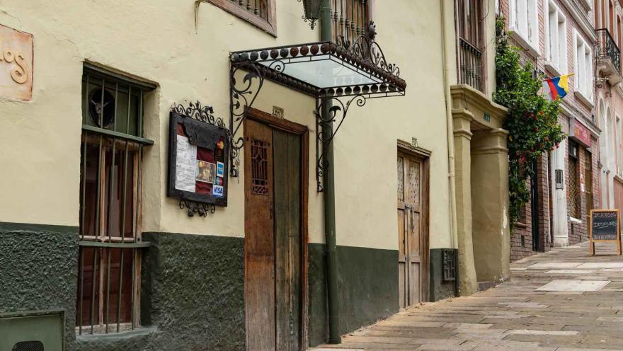 La fotografía muestra una calle adoquinada en La Candelaria. El enfoque principal está en un edificio de fachada ocre con un zócalo de color verde oscuro, que alberga lo que es una cafeteria. Una puerta de madera rústica y una ventana con rejas de hierro forjado resaltan el estilo colonial del lugar. 