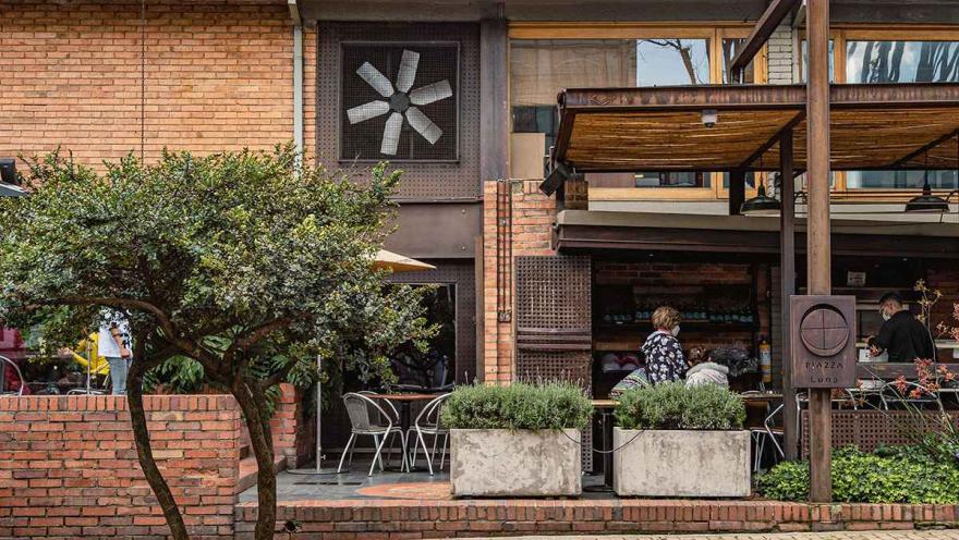 El edificio, con sus paredes de ladrillo y detalles de madera, tiene una gran ventana con un ventilador que le da un toque distintivo. Una terraza exterior, delimitada por jardineras de cemento con plantas verdes, invita a los comensales a disfrutar del aire libre. Los ladrillos de la acera y las mesas al aire libre contribuyen a la atmósfera relajada y moderna del lugar.