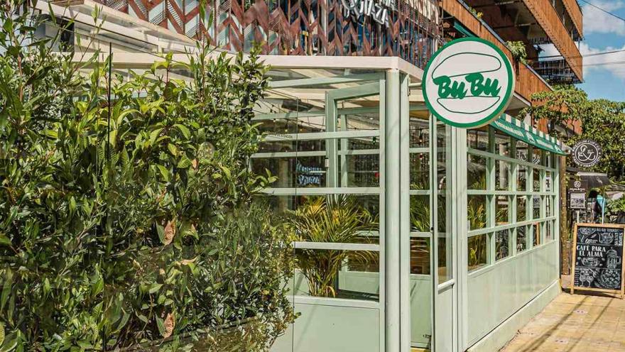 Esta foto de cerca muestra una parte de un café en el Parque de la 93. La estructura, que parece ser una extensión de un edificio, está hecha de vidrio y marcos de color verde claro, lo que le da un aspecto moderno y limpio. Un letrero redondo con el nombre "Bu Bu" y un icono de una hamburguesa de estilo minimalista cuelga en la parte superior. Alrededor de la estructura de vidrio, plantas verdes aportan frescura y privacidad.