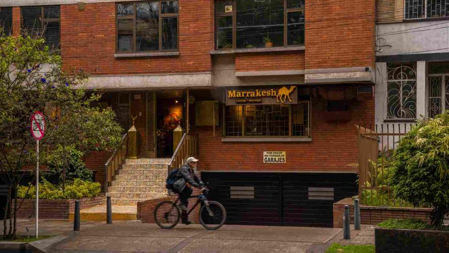 La fotografía muestra la entrada de 'Marrakech', un restaurante con una fachada de ladrillo rojo en el Park Way. La puerta principal está flanqueada por una escalera de azulejos y barandillas doradas, lo que le da un toque exótico. El letrero del restaurante, con el nombre en letras árabes y la silueta de un camello, evoca la cultura de Marruecos. 