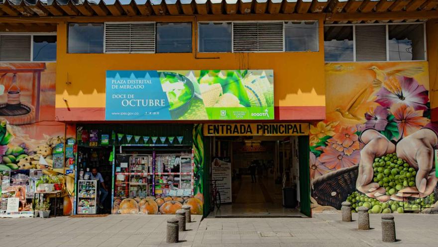  Esta fotografía presenta la fachada exterior de la "Plaza Distrital de Mercado Doce de Octubre". El edificio es de color amarillo brillante y cuenta con grandes murales vibrantes en ambos lados de la entrada principal, que representan frutas, verduras y manos sosteniendo guisantes. Un gran letrero azul con información del mercado se encuentra sobre la entrada. La "ENTRADA PRINCIPAL" está claramente marcada y abierta, revelando un pasillo interior.