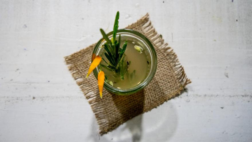 Esta es una vista cenital de un vaso pequeño con una bebida de color verde pálido, decorada con una ramita de romero y dos pétalos de naranja o cáscaras en forma de luna. El vaso está colocado sobre un pequeño trozo de arpillera cuadrada, que a su vez se encuentra sobre una superficie de madera blanca. La iluminación crea una sombra suave debajo del vaso y el posavasos.