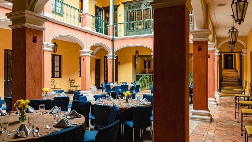 Esta imagen captura el impresionante patio central del Hotel de la Ópera, un lugar icónico en el centro de Bogotá. El espacio, de estilo colonial, presenta arcos y columnas en los dos pisos que lo rodean. Las paredes en tonos ocre, naranja y amarillo crean una atmósfera cálida y luminosa. Mesas redondas vestidas con manteles marrones y sillas azules están dispuestas para eventos o comidas, y en el centro, pequeños floreros con flores amarillas añaden un toque de color. 