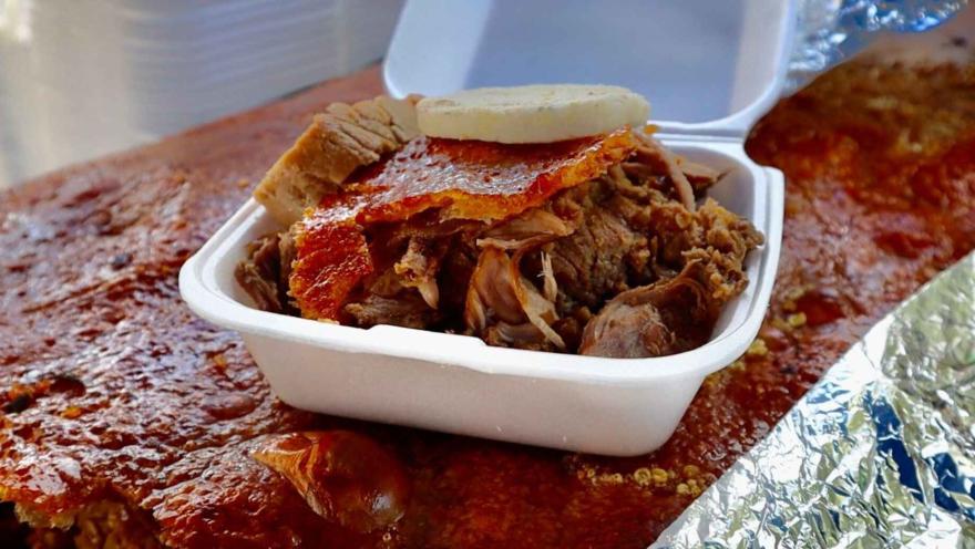Esta fotografía de cerca se enfoca en un plato de lechona servido en un recipiente blanco de icopor. La lechona, con su carne jugosa y tierna, está coronada con un trozo de la crujiente piel de cerdo y un pedazo de arepa. La textura de la carne desmenuzada y el color dorado de la piel son los protagonistas de la imagen, que evoca el sabor y la tradición de este plato típico colombiano. En el fondo, se ve el resto de la lechona entera, lista para ser disfrutada.