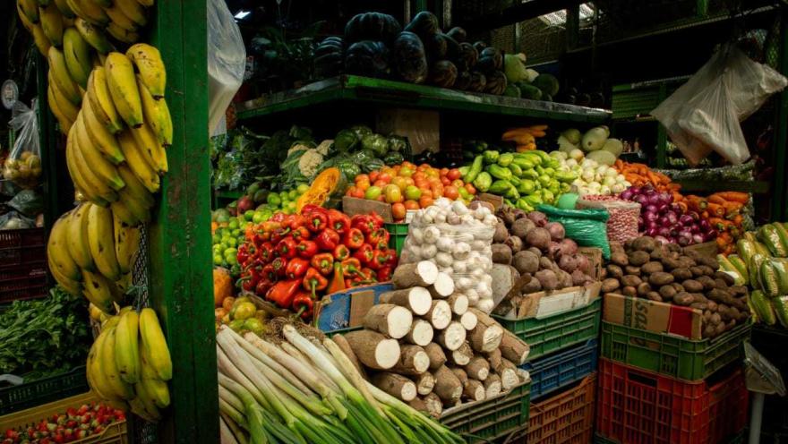 Un primer plano detallado de un puesto de frutas y verduras en el mercado, mostrando una impresionante variedad y abundancia de productos. Grandes racimos de plátanos amarillos cuelgan en el lado izquierdo. El puesto está densamente lleno de pimientos rojos, tomates pequeños, cebollas rojas, zanahorias, raíces de yuca apiladas, patatas, lechugas y otras hortalizas. Los productos están dispuestos en cestas y cajas, creando una explosión de colores y texturas.