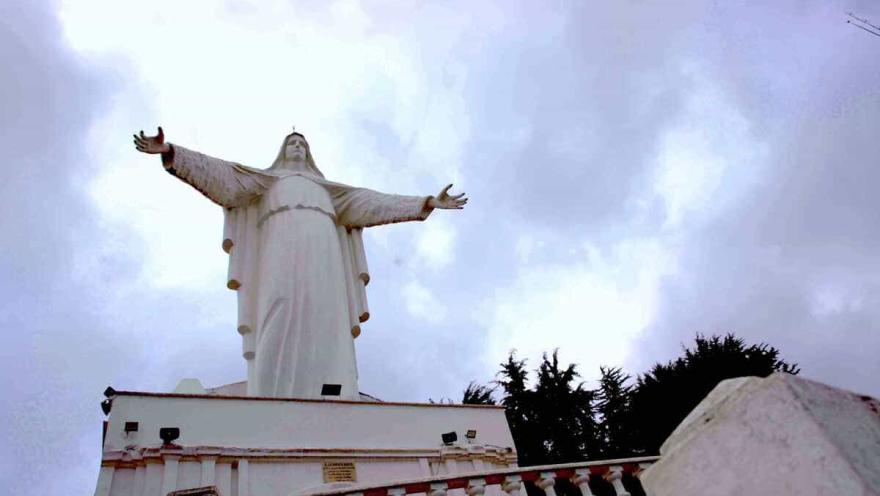 White statue of the Virgin of Guadalupe with open arms, above the church and under a blue sky with dark clouds. It is surrounded by tree tops.