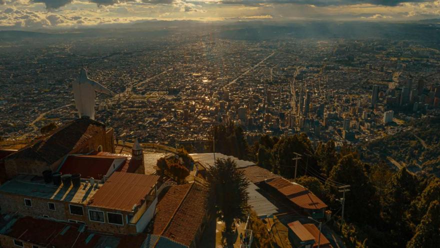 Guadalupe Church from above and the back of the white statue, surrounded by green trees, with Bogotá visible in the distance with a blue sky and white clouds.