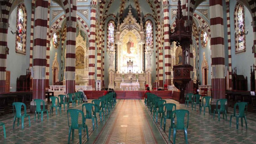 Santuario Nuestra Señora del Carmen en el interior con luz artificial destacan las columnas con franjas horizontales de color rojo y blanco, el púlpito en madera y al fondo el altar con una luz brillante sobre una imagen de la Virgen. Al centro de la iglesia hay cuatro filas de plástico vacías y dispuestas para los feligreses.