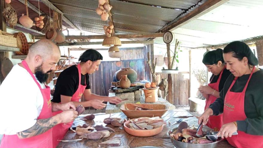 Dos hombres y dos mujeres con delantales rojos, cocinando mazorcas en una cocina artesanal.