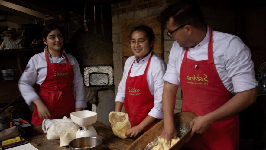 Dos mujeres jóvenes y un hombre jóven amasan amasijos en una cocina artesanal.