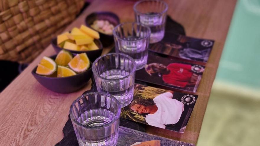 Cuatro copas pequeñas con viche sobre sendos portavasos con fotografías de personas de raza negra. En la mesa hay unas vasijas con limón, piña y maní.